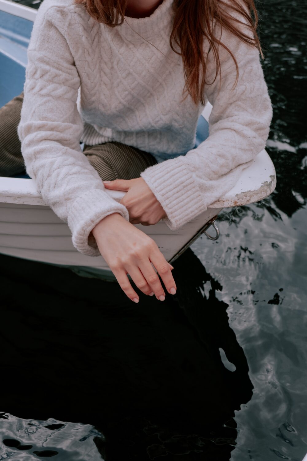 Person in cozy sweater sitting on boat, hand over water.
