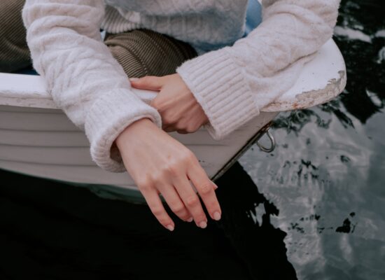 Person in cozy sweater sitting on boat, hand over water.