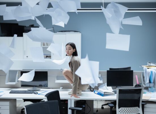 Person crouching on office desk, papers flying everywhere, chaotic scene.