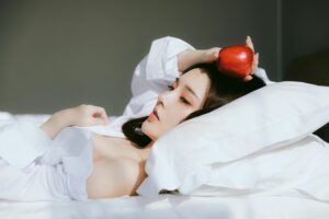 Person lying on bed, holding a red apple, white sheets.