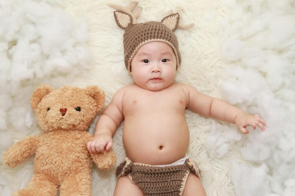 A baby wearing a deer costume with a teddy bear on a fluffy white rug.
