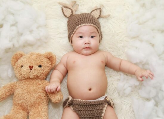 A baby wearing a deer costume with a teddy bear on a fluffy white rug.