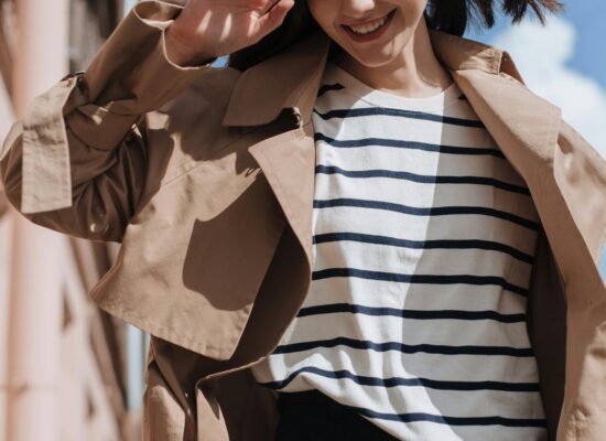 Person smiling outdoors, wearing trench coat and striped shirt.