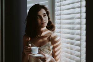 Person holding teacup, standing by window with blinds, thoughtful.