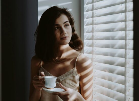 Person holding teacup, standing by window with blinds, thoughtful.
