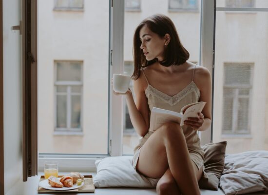 Person sitting by window, drinking coffee, reading book, breakfast nearby.