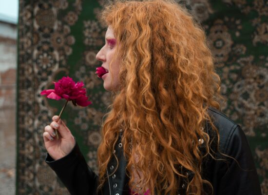 Person with red hair holds a pink flower to their mouth.