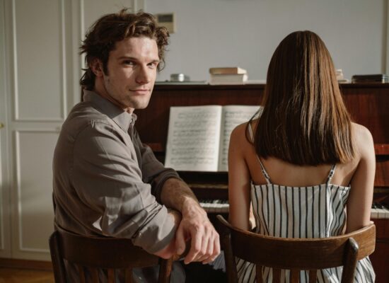 Two people sit at a piano with sheet music open.