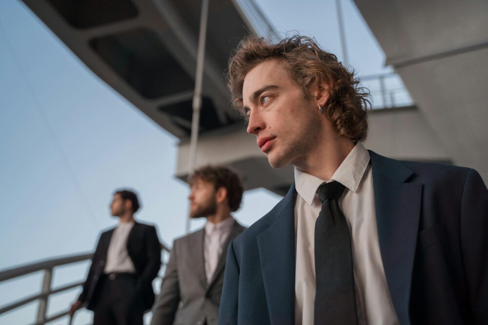 Three men in suits stand under a bridge looking out.