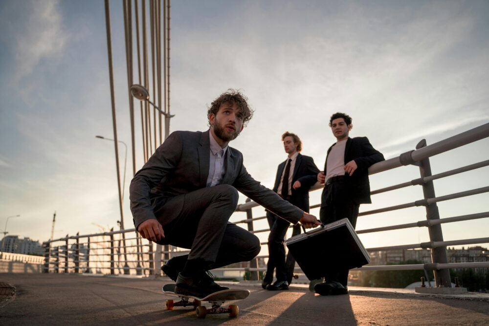 Three men in suits on a bridge with a skateboard and briefcase.
