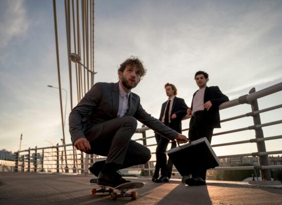 Three men in suits on a bridge with a skateboard and briefcase.