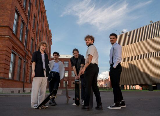 Four young men with skateboards stand outside buildings.