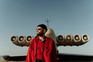 Person in red shirt stands before large, unusual aircraft outdoors.