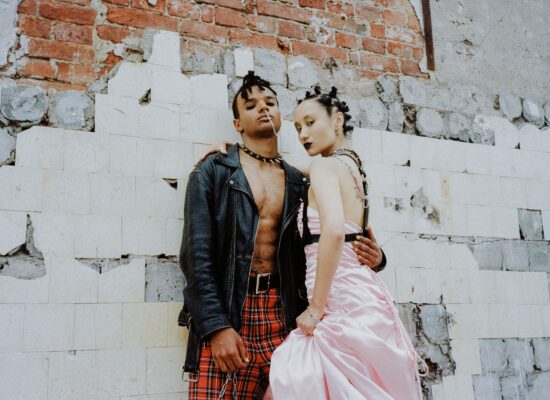 Two people in edgy fashion stand against a distressed brick wall.