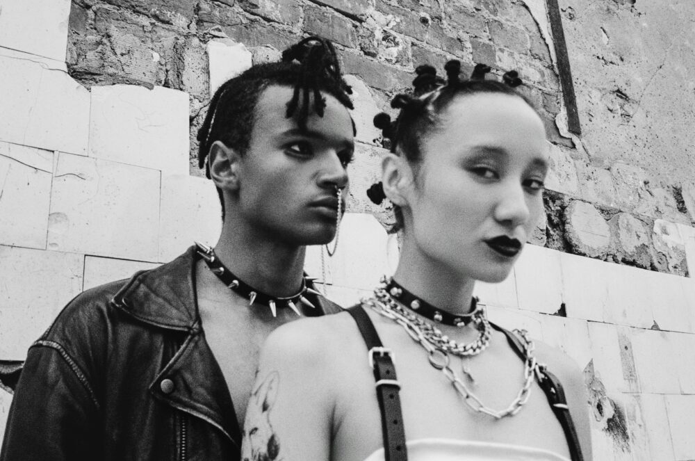 Two people with punk style accessories against a brick wall.