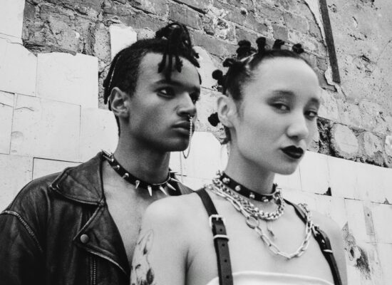 Two people with punk style accessories against a brick wall.