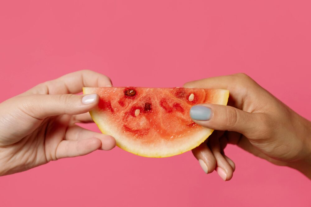 Two hands holding a watermelon slice against pink background.