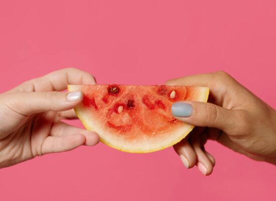 Two hands holding a watermelon slice against pink background.