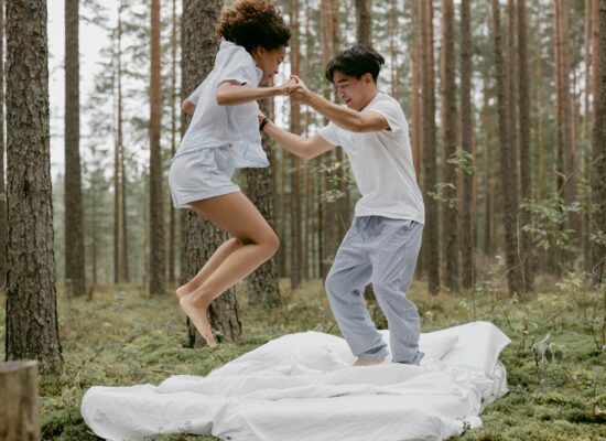 Two people jumping on a bed outdoors in a forest.