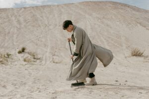 Person in long coat sweeping sand in a desert landscape.