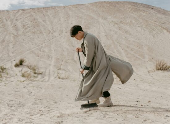 Person in long coat sweeping sand in a desert landscape.