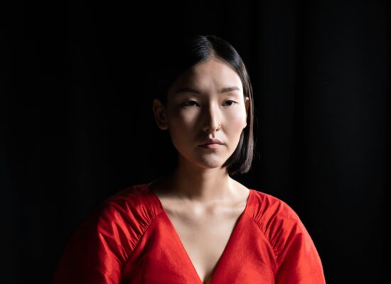 Person in red top against dark background, neutral facial expression.