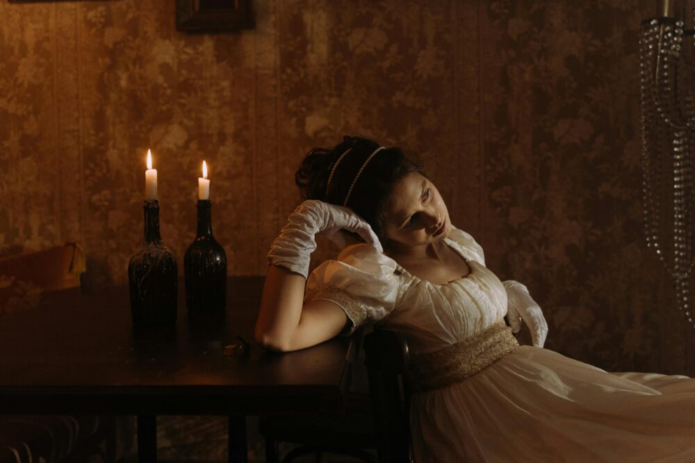 Person in vintage dress sits by candlelit table, looking contemplative.