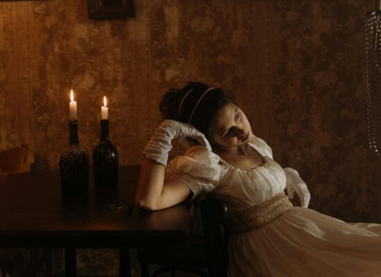Person in vintage dress sits by candlelit table, looking contemplative.