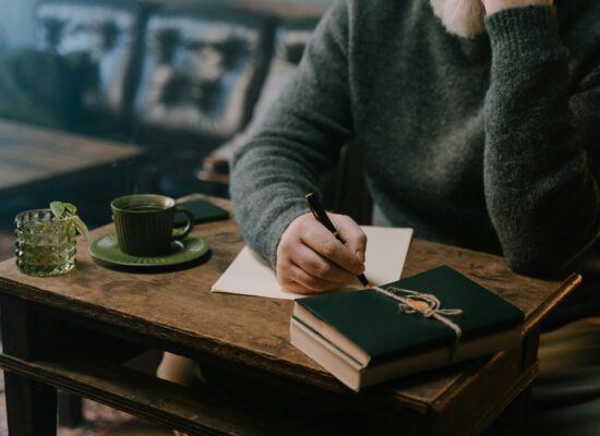 Person writing at a wooden table with drinks and books.
