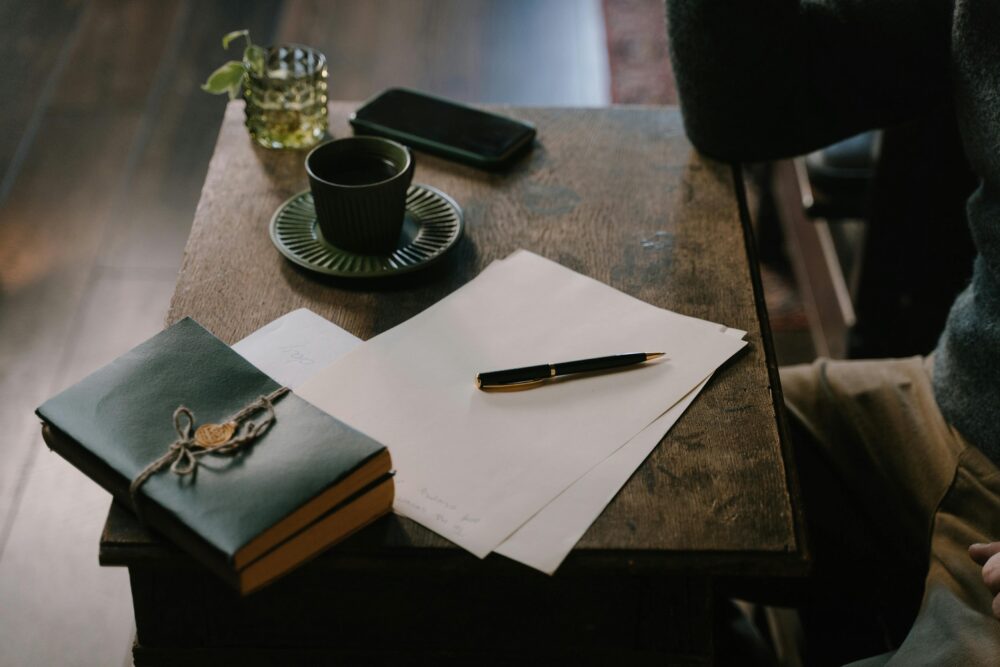 Notebook, pen, papers, coffee, and phone on wooden table.