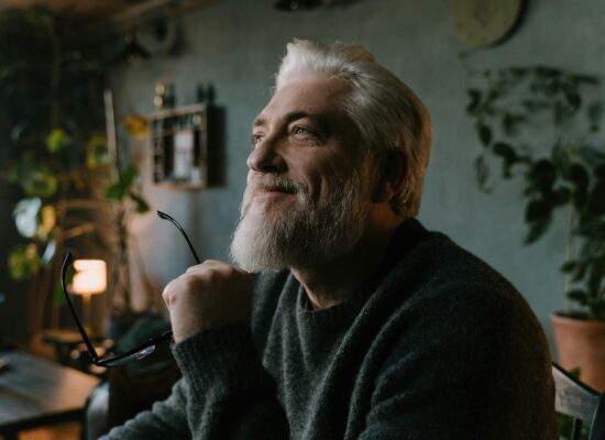Person smiling thoughtfully indoors, holding glasses, surrounded by plants.