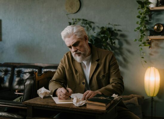 Person writing at desk, surrounded by books and crumpled paper.