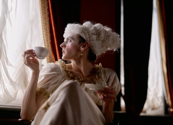 Person in historical costume drinking tea by elegant window curtains.