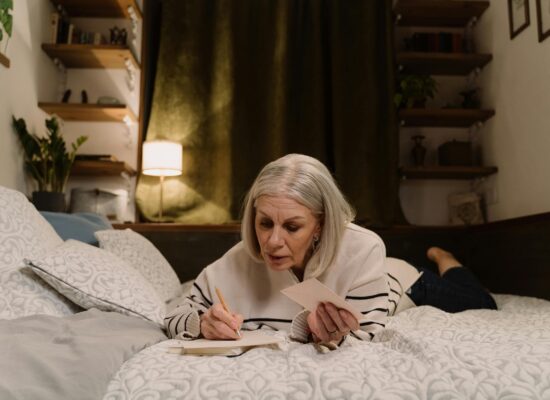 Person lying on bed, writing in notebook, holding envelope, cozy room.