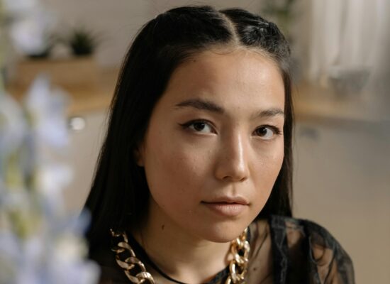 Person with long dark hair and gold chain necklace indoors.