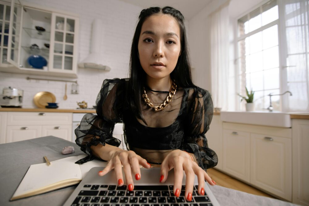 Person typing on laptop in bright kitchen, notebook nearby.