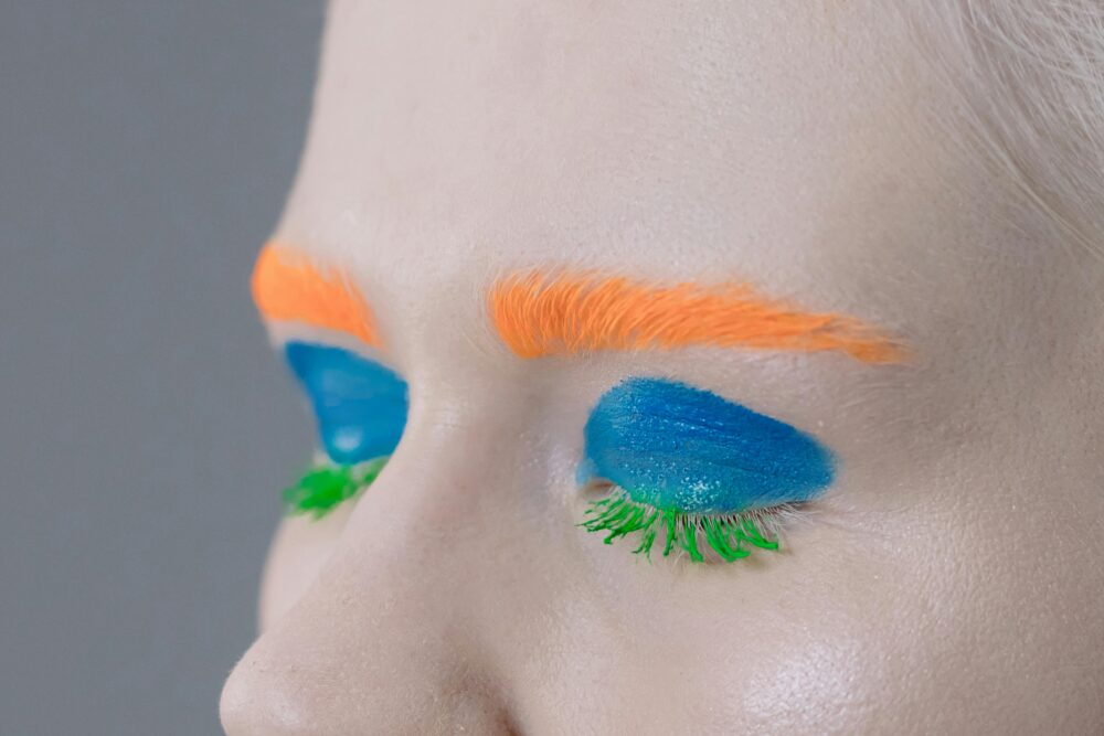 Bright orange eyebrows, blue eyeshadow, and green mascara on eyelashes.