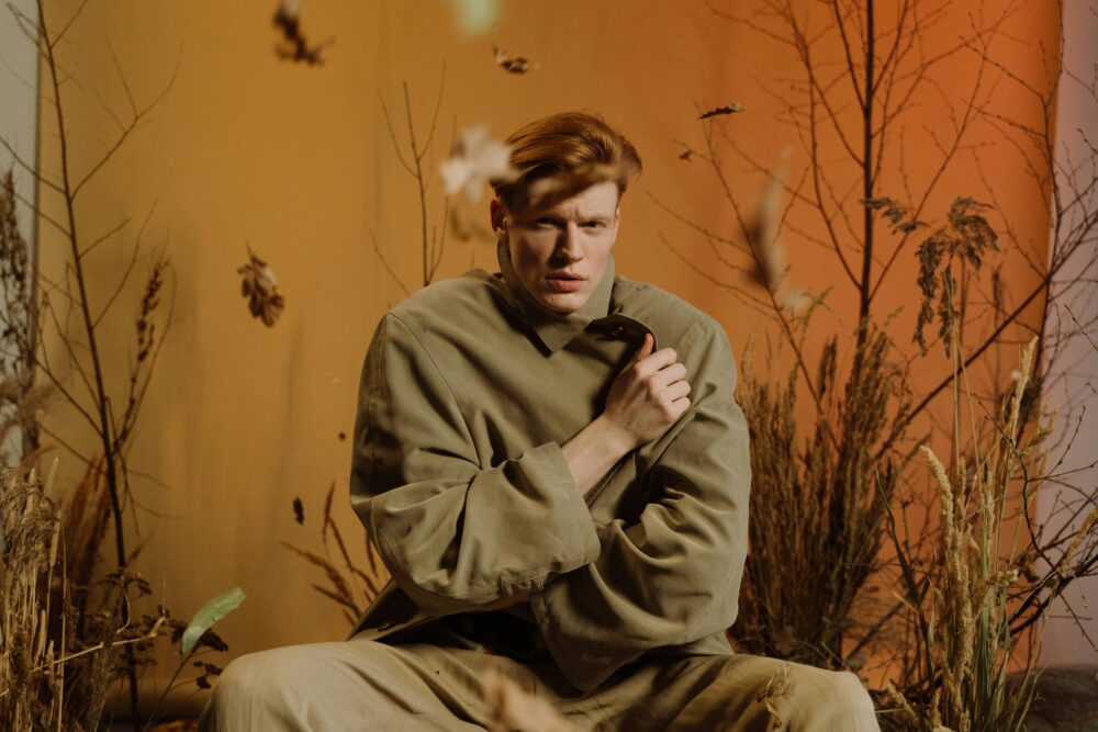 Person in oversized coat surrounded by dry autumn plants indoors.
