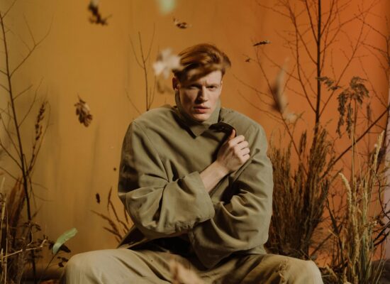 Person in oversized coat surrounded by dry autumn plants indoors.