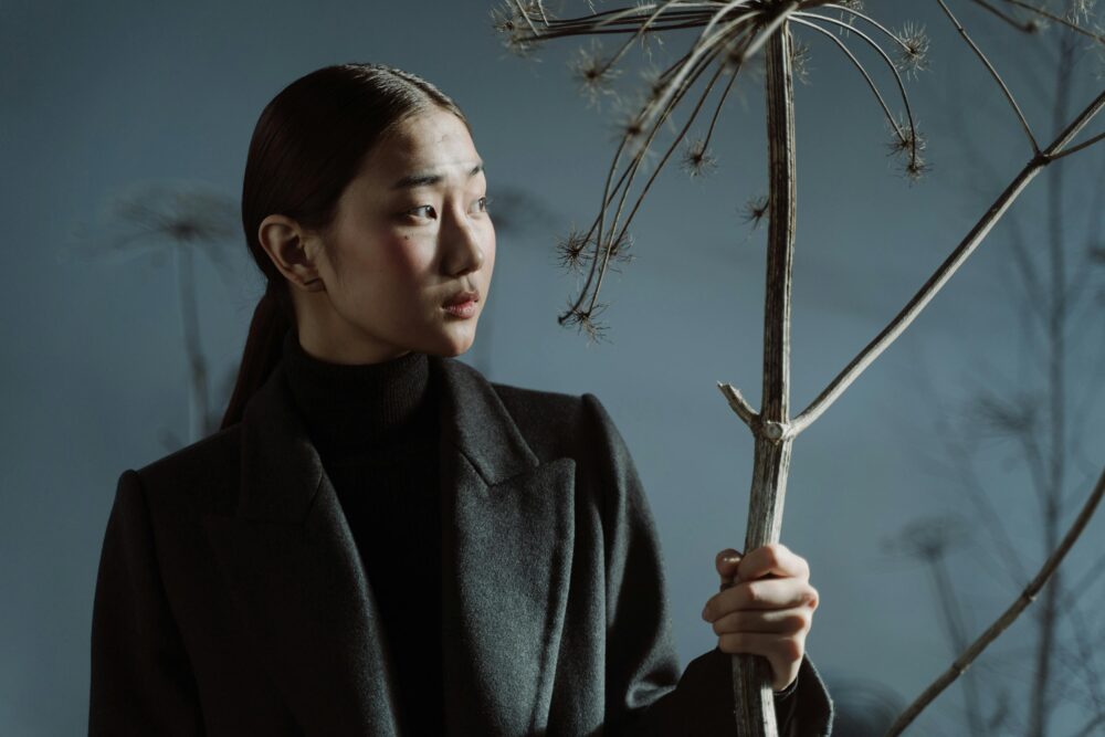 Person in dark coat holding dried plant, looking sideways thoughtfully.