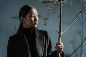 Person in dark coat holding dried plant, looking sideways thoughtfully.