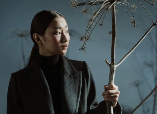Person in dark coat holding dried plant, looking sideways thoughtfully.