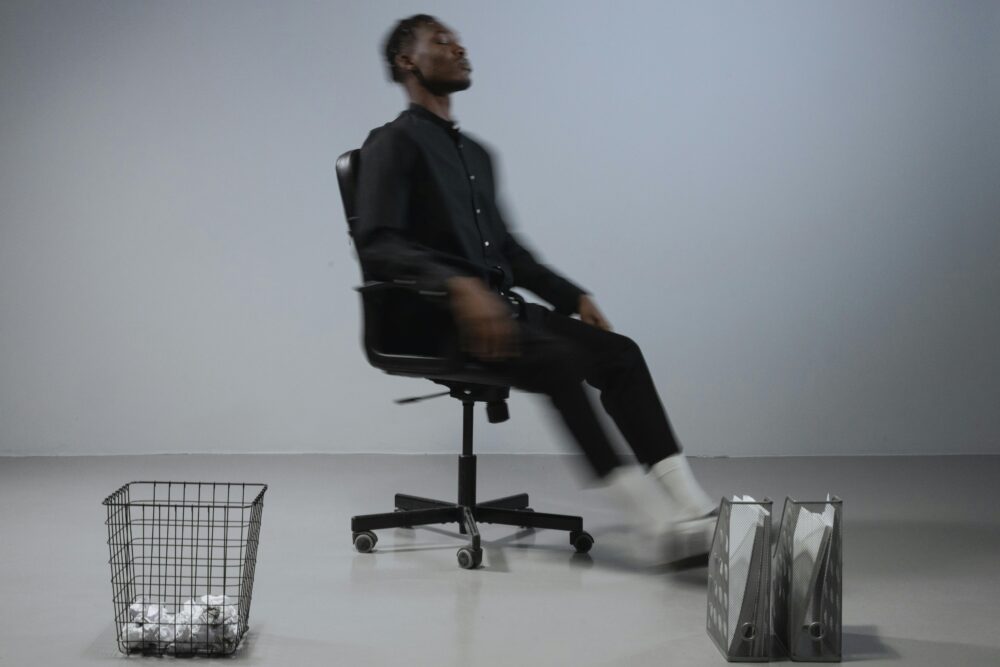 Person in black clothing spinning in an office chair with a trash can.