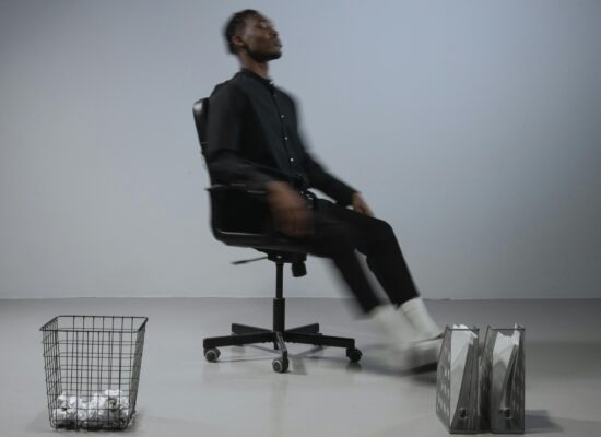 Person in black clothing spinning in an office chair with a trash can.