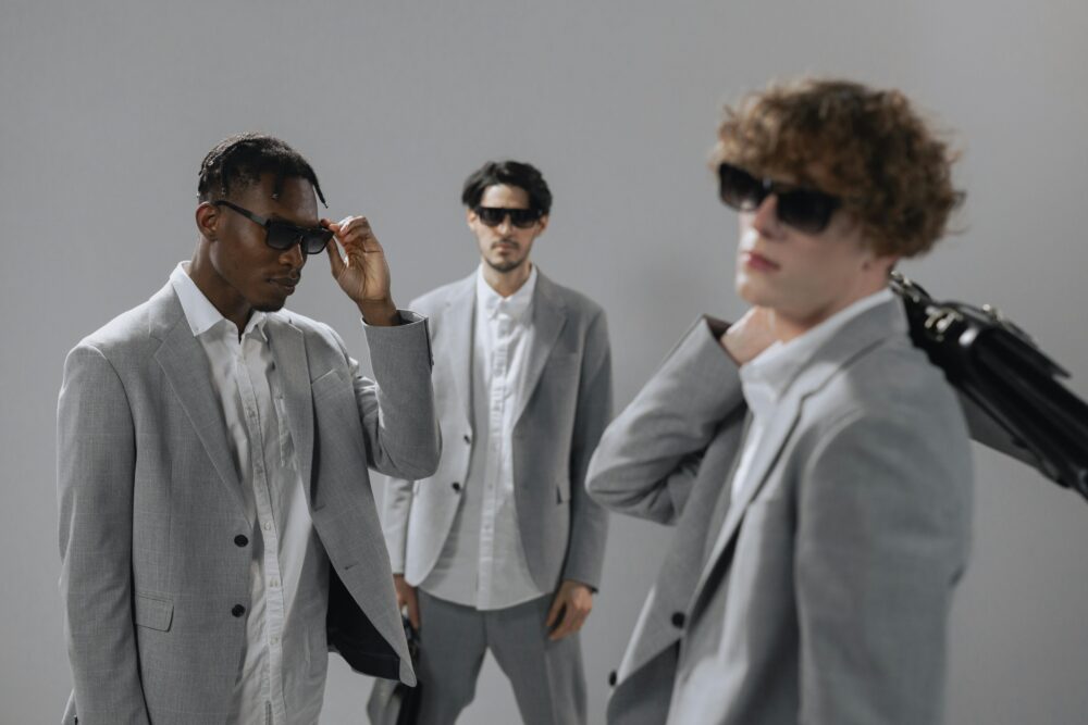 Three men in grey suits and sunglasses pose against a grey background.