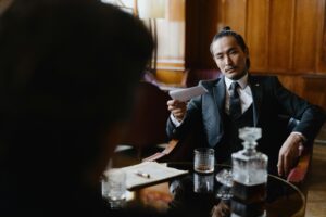 Man in suit offers paper, whiskey decanter and glass on table.