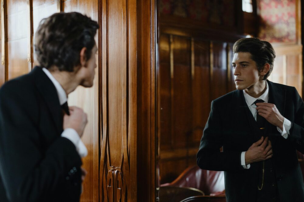 Man in a suit adjusts his tie in a mirror.