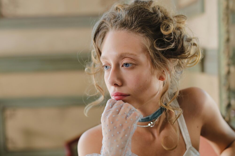 A woman with blue eyes and blonde hair wears a choker and gloves.