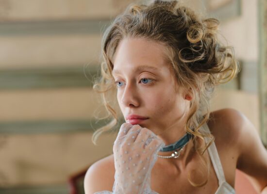 A woman with blue eyes and blonde hair wears a choker and gloves.