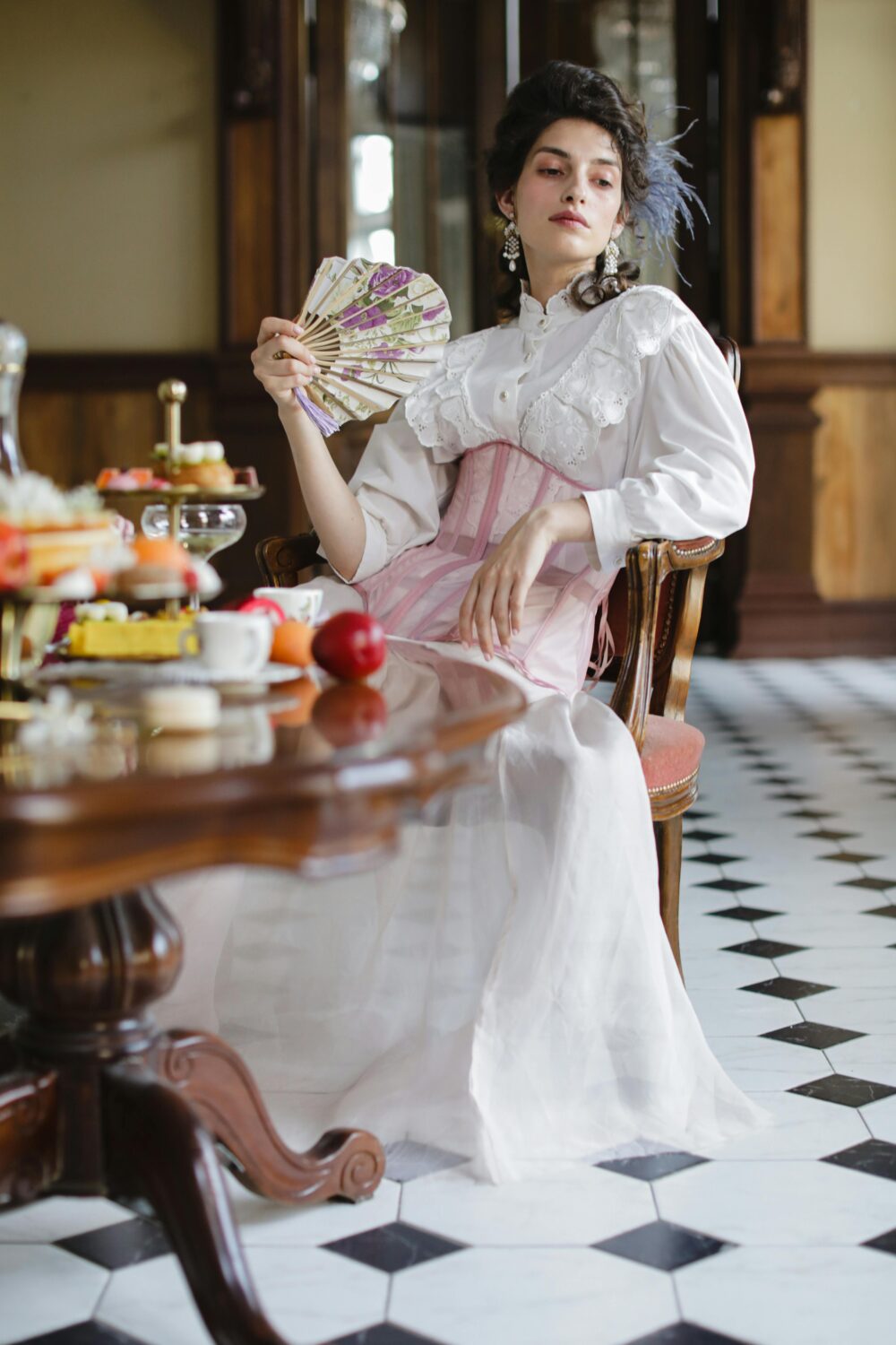 A person in vintage attire holds a fan near a table with desserts.